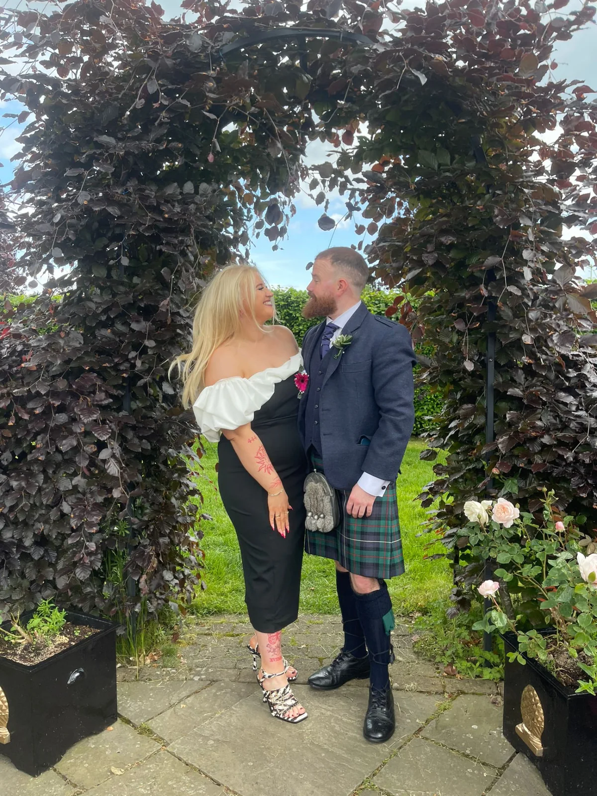 Holly and John smiling together beneath a leafy garden arch